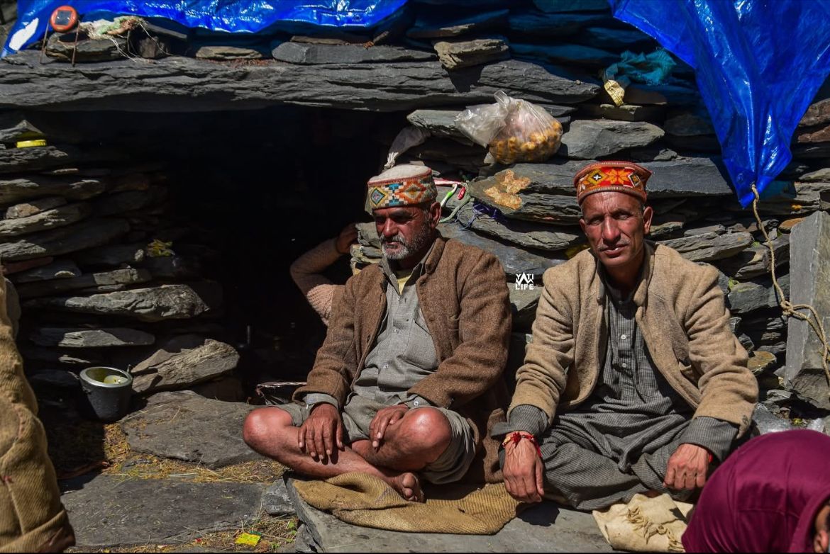 Two Gaddi elders in traditional Chambyali caps seated outside a slate-stone shepherd hut in Saal Valley, Chamba